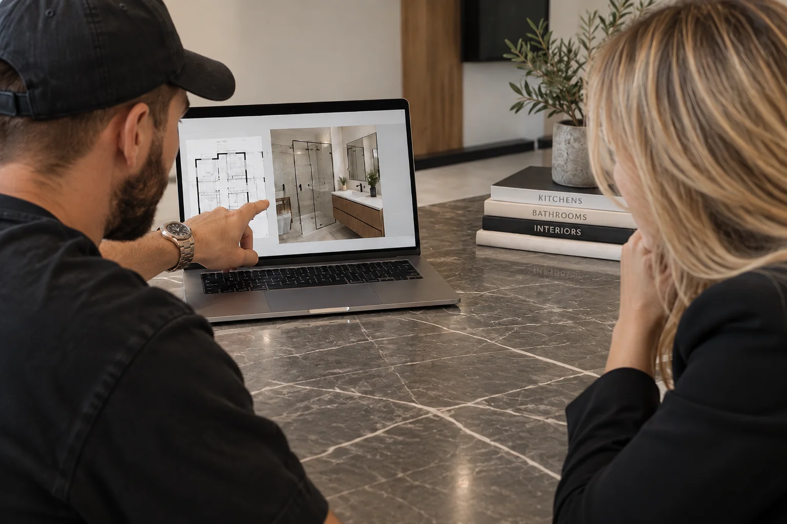 Contractor and client reviewing bathroom 3D render and floor plan on laptop at marble table with Kitchens Bathrooms Interiors books